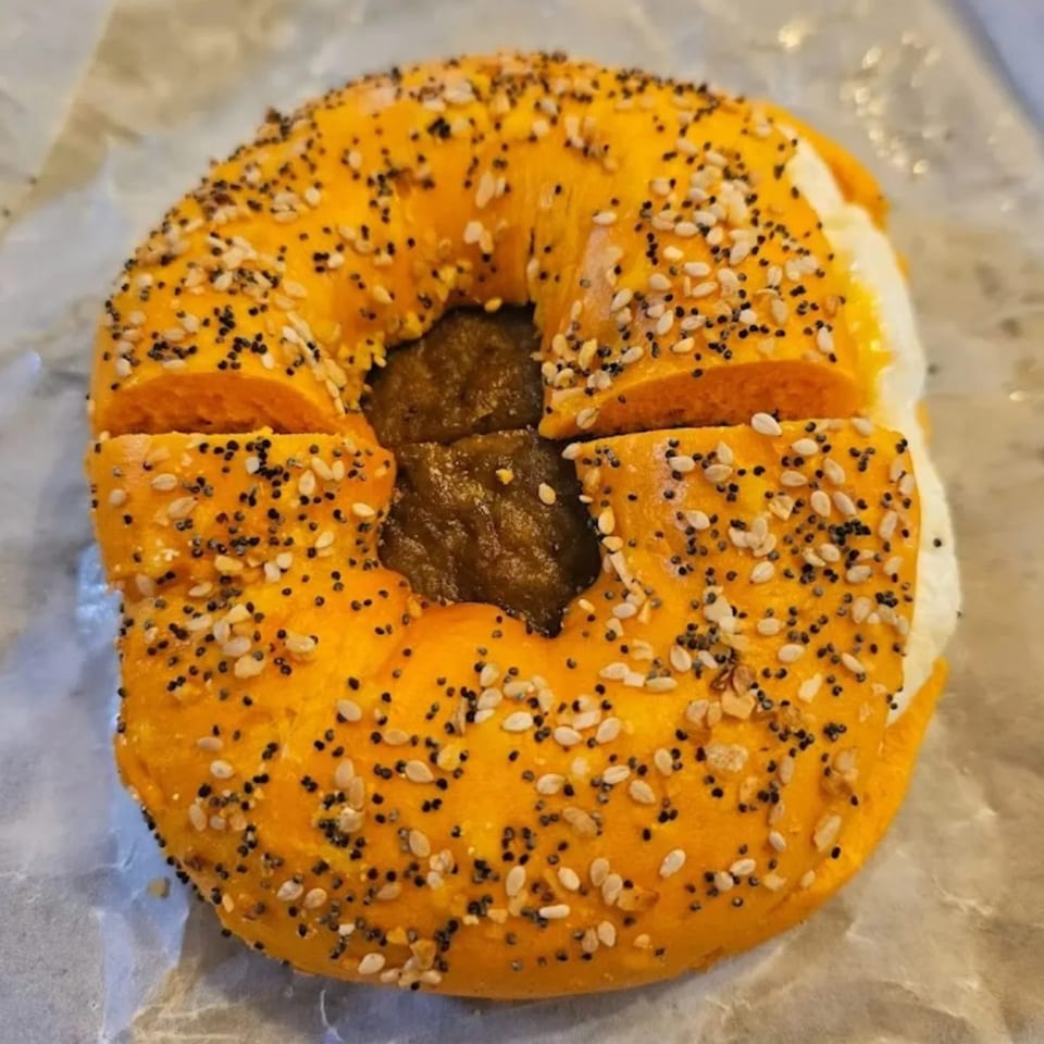 A golden bagel from Bagel & Deli Creations, topped with poppy and sesame seeds, filled with cream cheese and a dark spread.